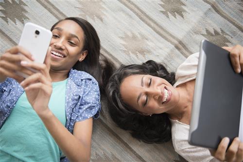 An African-American mother and daughter lay smiling on the floor. The daughter is looking at her phone and the mother her tab