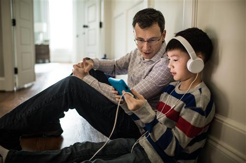 A father sits in the floor next to his son who has on headphones and s tablet