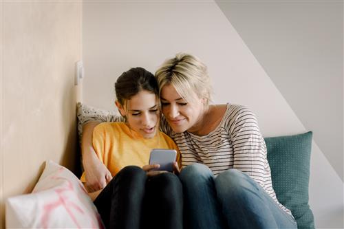 A blonde mother sits on a pillow with her teen daughter, both are looking at their phones