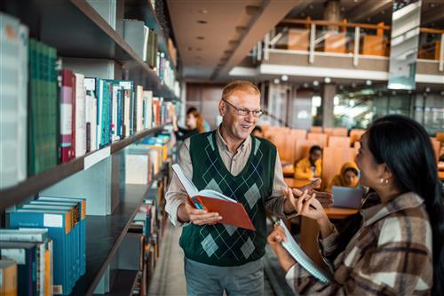 An older, caucasian teacher talks to a student in a library.