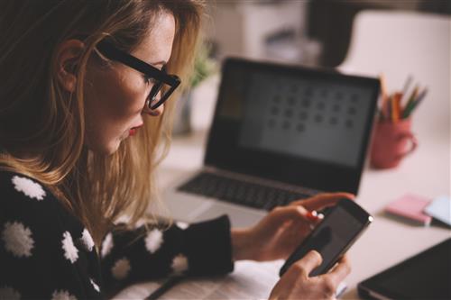 A woman with glasses stares at her phone. In the background is a laptop.