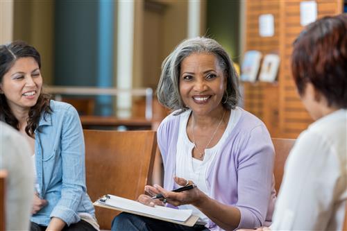 An African American woman with grey hair sits beside a dark-haired woman. On the woman's lap is a clipboard.